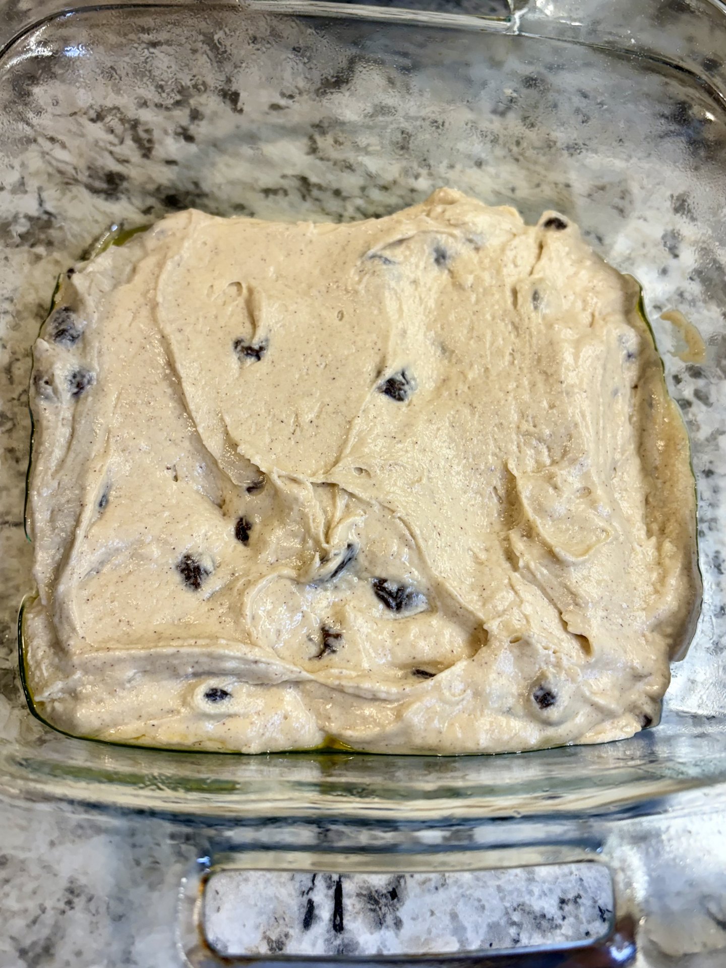 Pale coffee cake batter spread in a buttered glass baking dish, raisins evenly distributed throughout