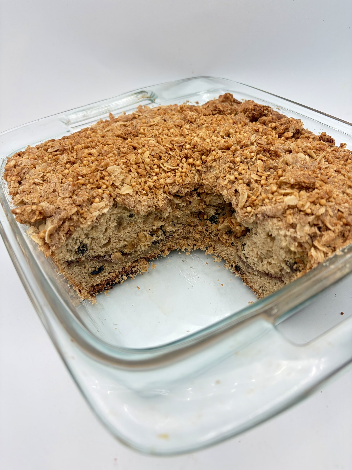 Granola coffee cake in the pan with a corner cut out, golden topping intact and the cinnamon ribbon visible through the crumb