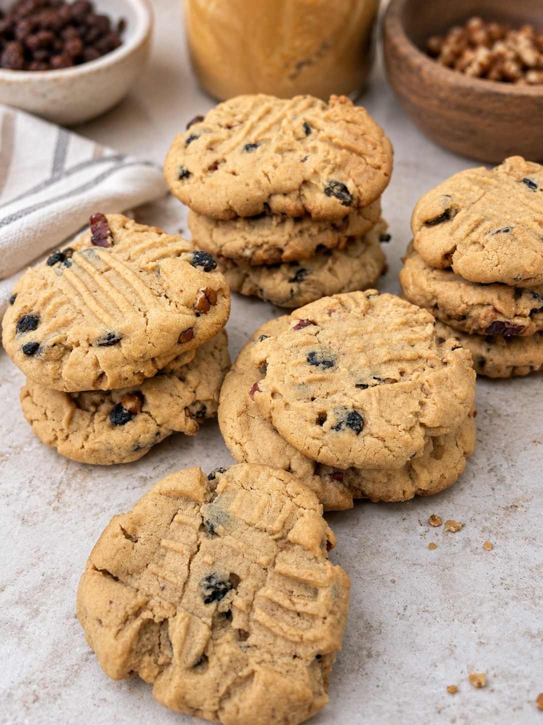 Peanut butter raisin cookies stacked on a counter, studded with raisins and walnuts, with a jar of peanut butter and bowls of raisins and walnuts behind them