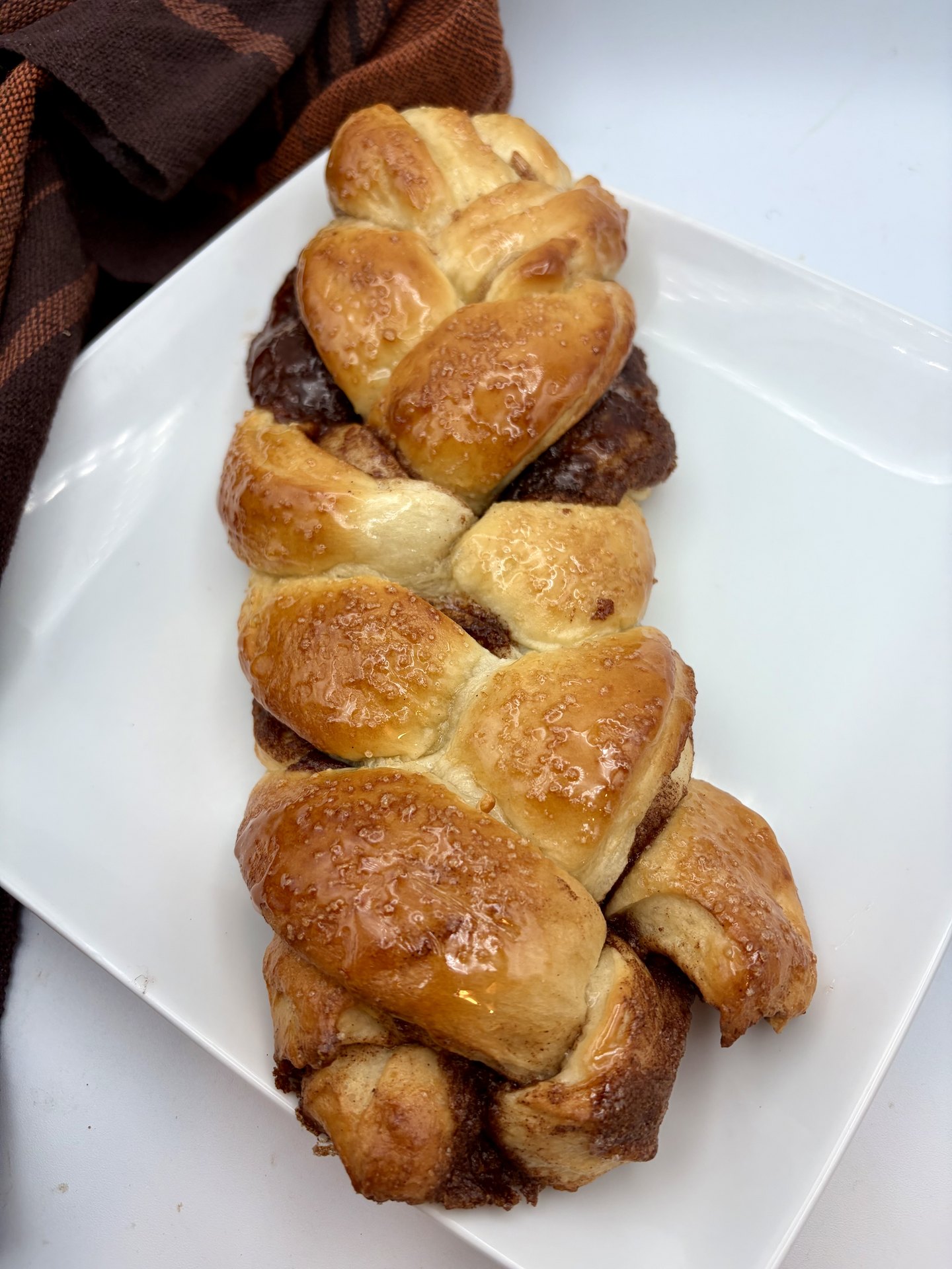The finished braided loaf, deeply glazed and shining, on a white square plate beside a rust colored towel