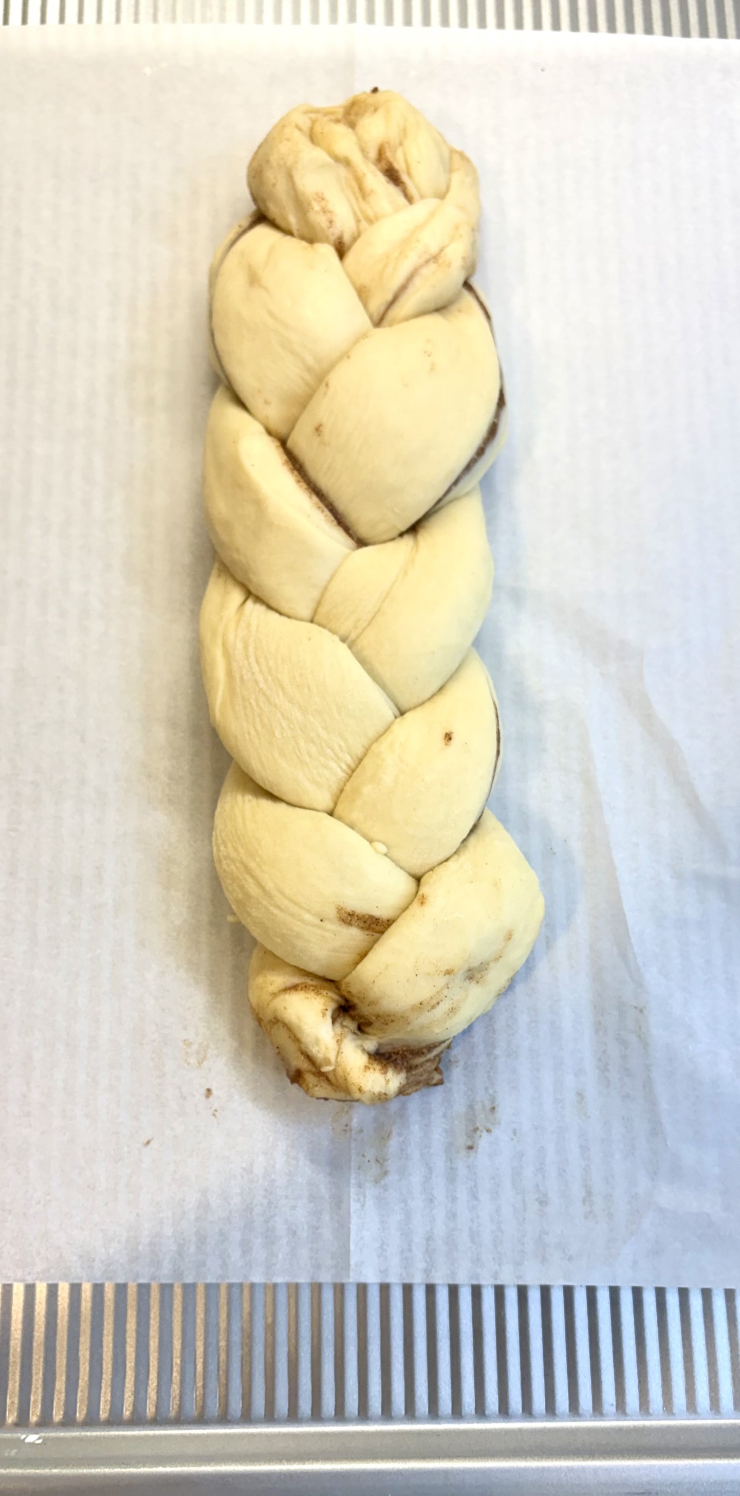 An unbaked four-strand braided loaf on parchment on a sheet pan, cinnamon filling showing between the strands