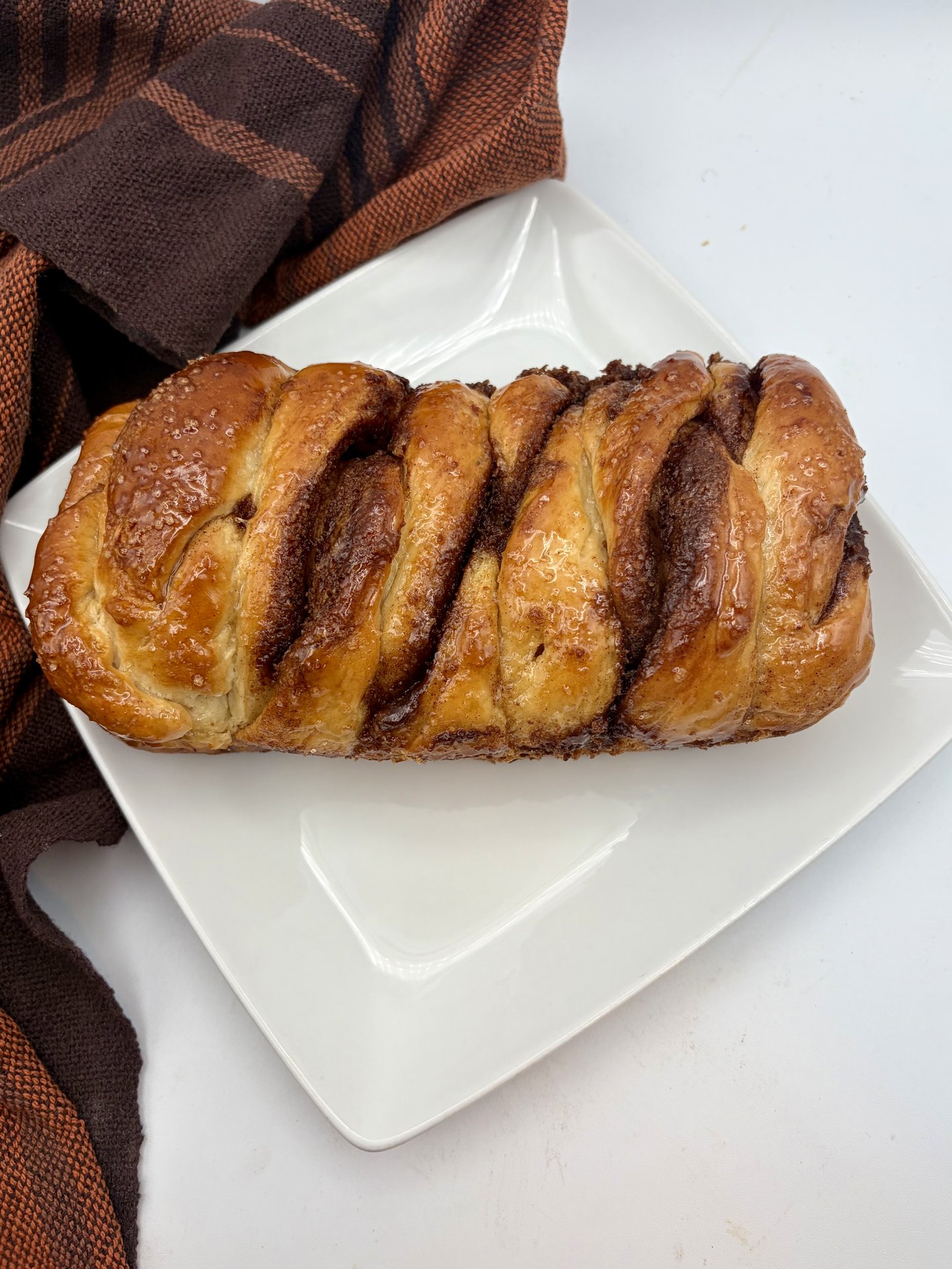 A glazed cinnamon accordion pull-apart loaf on a white square plate beside a rust and brown kitchen towel