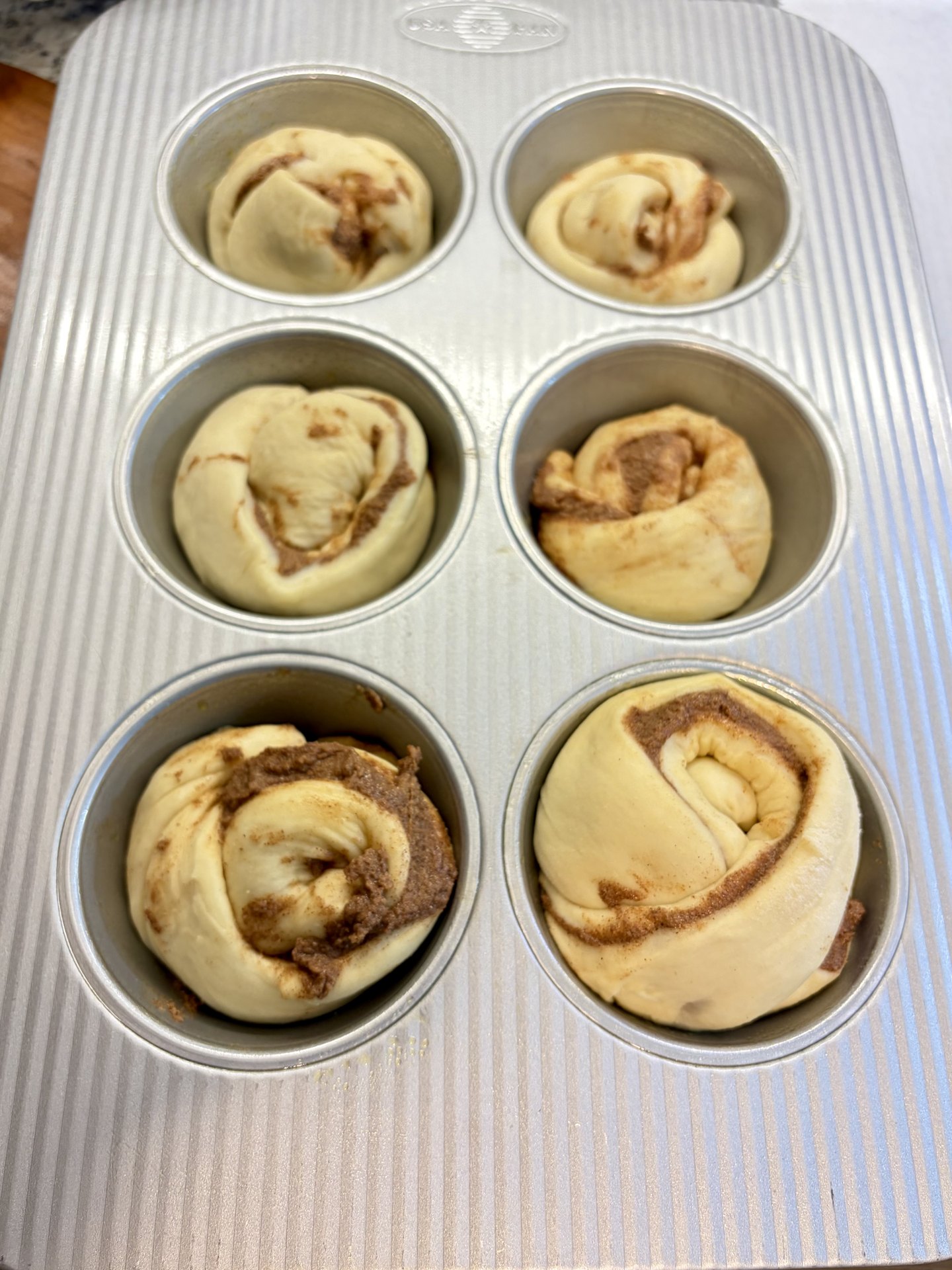 Six shaped cinnamon rolls sitting in a muffin pan before proofing, the filling visible in each swirl